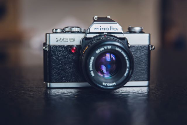 This image depicts a vintage Minolta XG9 camera placed on a dark surface. The camera shows classic styling with a reflective lens, highlighting the old-fashioned charm of film photography gear. Perfect for use in articles about the history of photography, retro photography enthusiasts, or as a nostalgic visual in a variety of photography-related content.