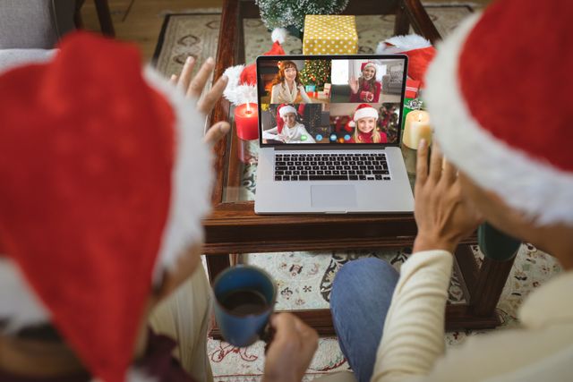 Couple wearing Santa hats engaging in a cheerful video call with friends displayed on a laptop. Hands waving to friends on the screen. Ideal for use in campaigns promoting virtual holiday gatherings, festive online interactions, or products supporting digital communication during holidays.