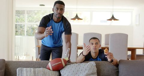Diverse friends watching football at home reacting and reaching over sofa