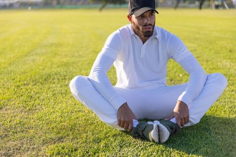 Cricket player resting cross-legged on sunlit grassy field in white kit and black cap