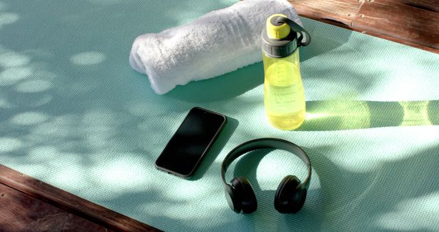 A rolled white towel, yellow water bottle with a green cap, black headphones, and smartphone resting on a light blue yoga mat. The sunlight creates natural shadows on the mat, suggesting an outdoor setting. Ideal for use in promotional materials for fitness classes, exercise blogs, health and wellness websites, or advertisements for workout gear.