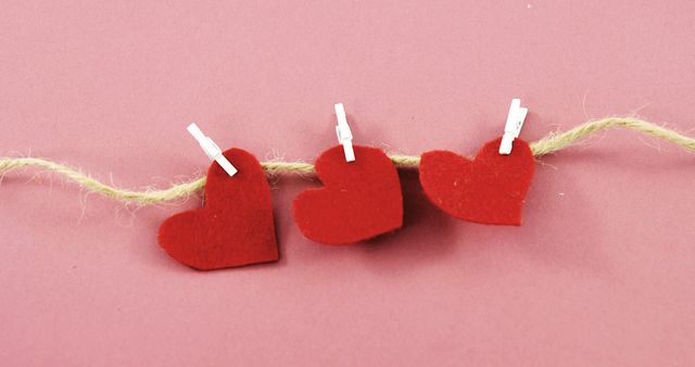Three red felt hearts are clipped to a twine string against a pink background, with copy space. Symbolizing love and affection, the image is often associated with Valentine's Day and romantic celebrations.