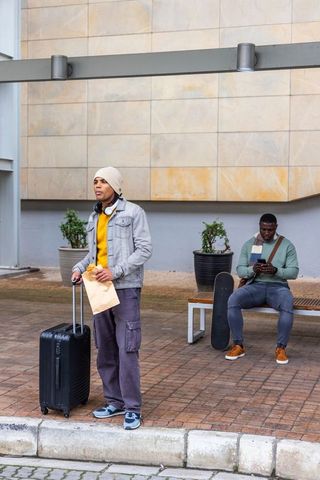 Standing traveler holding rolling suitcase and paper bag waiting at urban curb