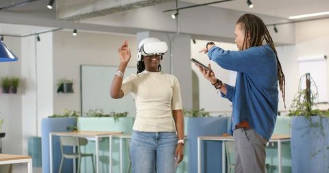 African American woman testing VR headset while colleague guiding via tablet in modern coworking hub