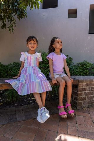 Two girls sitting on bench in sunlit courtyard wearing pastel striped dress and bright sneakers