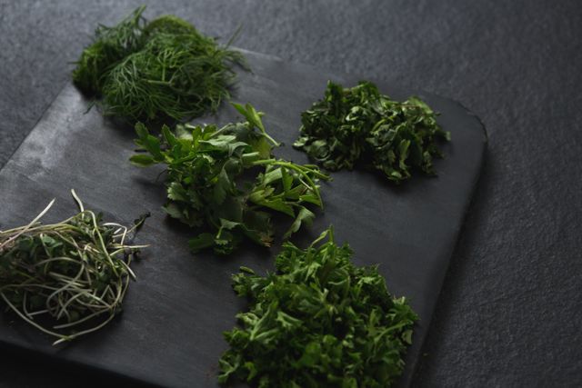 This image shows a close-up of various fresh herbs, including parsley, dill, cilantro, and microgreens, arranged on a dark chopping board. Ideal for use in culinary blogs, recipe websites, cooking magazines, and health food promotions. Perfect for illustrating articles on healthy eating, organic ingredients, and food preparation.
