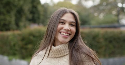 Smiling young woman wearing beige knit sweater posing outdoors with soft bokeh background