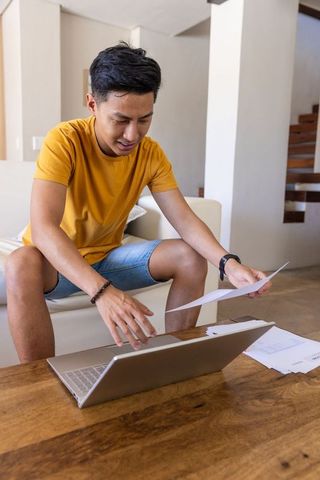 Asian man reviewing documents with laptop in living room workspace for remote work