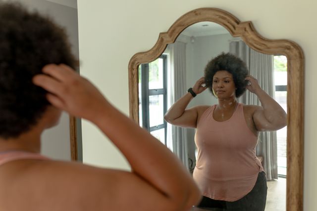 African american mid adult woman making hair while standing in front of mirror at home. unaltered, preparation, reflection, fitness and active lifestyle concept.