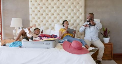 African american family unpacking suitcase on bed using tablet and smartphones smiling