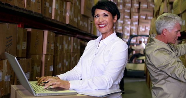 Businesswoman using laptop while standing in a warehouse with another worker. Ideal for business, logistics, supply chain management, e-commerce, and teamwork themes.