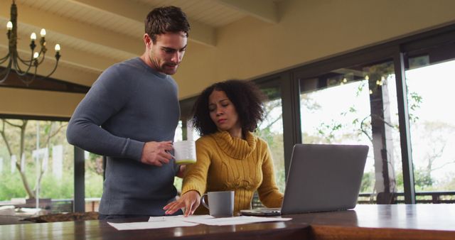 Couple working together in a cozy home office environment surrounded by nature. They are dressed in casual autumn attire, using a laptop while drinking coffee, suggesting a productive yet relaxed remote work setting. Ideal for illustrating work-from-home trends, teamwork in remote environments, or the balance of work and personal life.