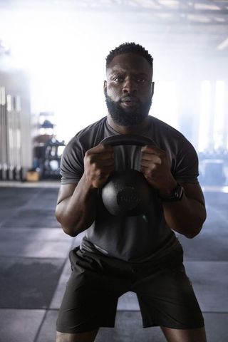 Powerful african american man squatting with kettlebell at chest in backlit gym