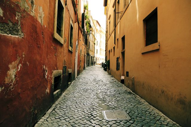 Narrow cobblestone street between aged buildings in an ancient European city offers a glimpse into historical architecture. Warm sunlight enhances the rustic charm. Ideal for travel blogs, heritage tourism promotions, and urban exploration themes.