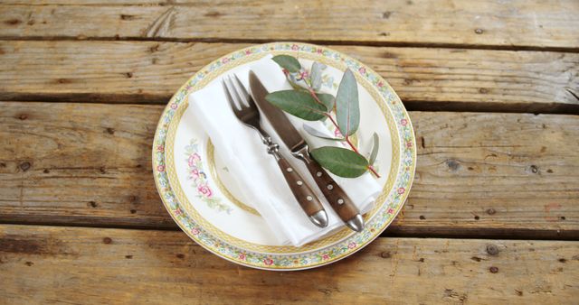 A vintage plate with floral patterns holds a neatly folded napkin, a fork, and a knife, accented with a sprig of greenery, all set on a rustic wooden table. This setting evokes a sense of homely elegance, perfect for a quaint dining experience or a themed event.