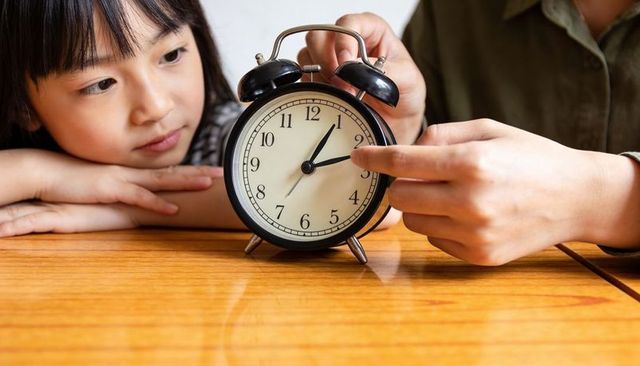 Child learning to tell time with classic black twin-bell alarm clock on wooden table