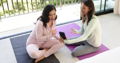 Diverse women friends sharing fitness app on phone while sitting on yoga mats on balcony