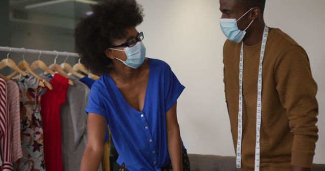 Image depicts two African American fashion designers collaborating on a new clothing collection while wearing face masks, highlighting pandemic safety measures. Suitable for use in articles or blogs on fashion industry adaptations during the pandemic, teamwork in creative professions, or showcasing diverse professionals in fashion design studios.