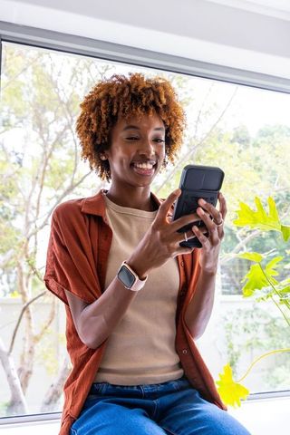 African american woman smiling while using smartphone and smartwatch on sunny windowsill