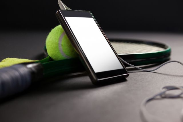 Close up of mobile phone and headphones by tennis ball with racket on black background