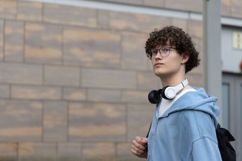 Curly-haired teen standing on urban sidewalk wearing blue hoodie and headphones