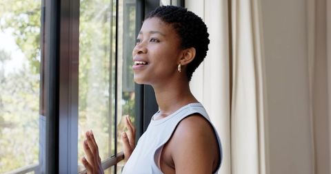 African woman looking out window touching glass, standing by balcony in morning light