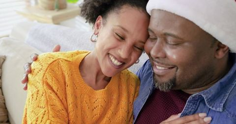 Sharing cozy holiday hug: Diverse couple smiling on sofa wearing Santa hat and sweater