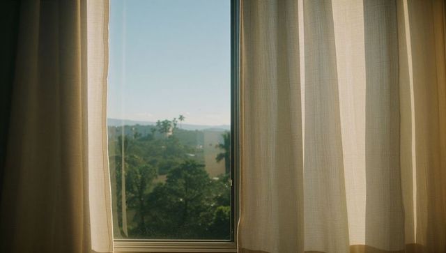 Sunlight streaming through beige curtains framing window with distant palm trees