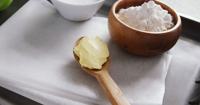 Image features essential baking ingredients: a wooden spoon with butter, a wooden bowl with flour, and a small bowl of granulated sugar. The items are arranged on parchment paper in a kitchen setting. Ideal for use in baking blogs, recipe websites, or cookbooks.