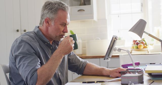 The scene shows a senior man at his home office, working on a computer while drinking coffee. Perfect for use in articles about remote work, work-from-home setups, or lifestyle choices of mature adults. Useful for illustrating the concept of productive home workspaces.