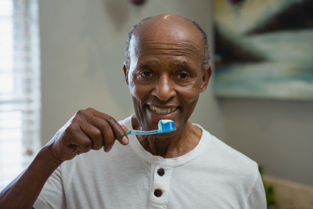 Senior man brushing teeth in bathroom at home, smiling. Ideal for promoting dental care, healthy lifestyle, and personal hygiene products. Useful for healthcare campaigns targeting elderly oral health.