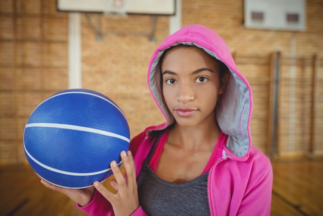 High school girl standing in gym holding basketball, wearing pink hoodie. Ideal for use in sports, youth empowerment, fitness, and educational content. Perfect for promoting athletic programs, school sports events, and motivational materials.