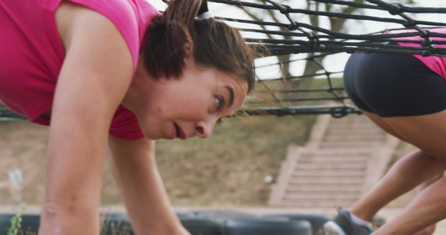 The woman is crawling under a net, participating in a fitness obstacle course. This can be used in materials promoting outdoor fitness challenges, teamwork activities, health and fitness events, or motivational content about physical endurance and determination.