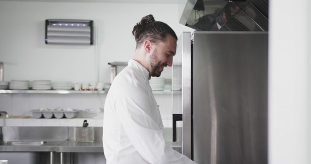 Professional chef dressed in white uniform standing in kitchen, opening oven door with a smile. Ideal for content related to gastronomy, cooking tutorials, culinary schools, restaurant advertising, or professional kitchen equipment.