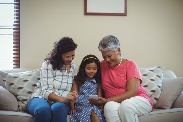 Three generations of women, including a grandmother, mother, and daughter, are sitting on a couch in a living room, using a mobile phone together. They are smiling and appear to be enjoying their time together. This image can be used for themes related to family bonding, technology use across generations, and home life. It is suitable for advertisements, blogs, and articles focusing on family, technology, and intergenerational relationships.