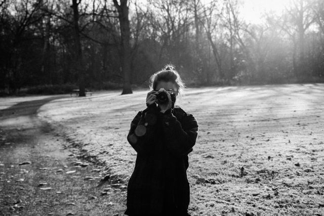 Individual holding camera, standing on frosty path in park with bare trees and early morning light. Perfect for themes like photography hobby, outdoor activities, winter mornings, and nature appreciation.