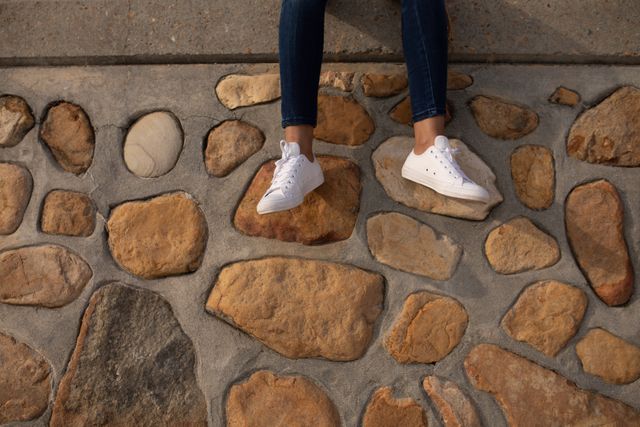 This image captures a young woman sitting on a stone wall, showcasing her casual style with white sneakers and blue jeans. Ideal for use in fashion blogs, lifestyle magazines, or advertisements promoting casual wear and outdoor activities. It can also be used for social media posts about fashion trends, relaxation, and enjoying nature.