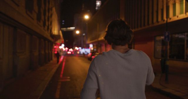 Individual seen from the back walking alone on an empty, dimly lit urban street at night. Streetlights and building façades create a moody, atmospheric vibe. Perfect for themes relating to city life, solitude, thoughtful moments, and urban exploration.