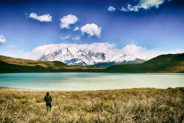 Traveler is standing and enjoying a panoramic view of a lake and snow-capped mountains with grassy landscape in the foreground. Perfect for themes related to travel, adventure, nature, hiking, exploration, and outdoor activities.