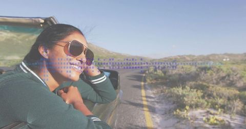 Smiling woman leaning out car window wearing aviator sunglasses and jacket with blue graphic overlay