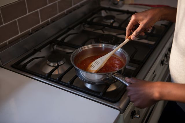 Mid section of woman stirring sauce in pot on gas stove in home kitchen. Ideal for use in articles or advertisements related to home cooking, culinary skills, kitchen appliances, or domestic life.