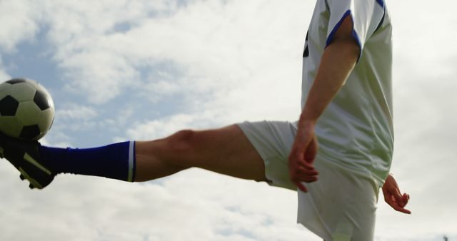 A soccer player in a white and blue uniform is about to kick a soccer ball, with copy space. Captured against a cloudy sky, the action shot emphasizes the dynamic movement and energy of the sport.