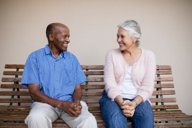 Smiling senior man and woman looking at each other while sitting on bench against wall - Download Free Stock Photos Pikwizard.com