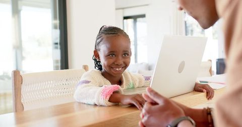 Smiling african american girl learning with guardian using laptop at home dining table