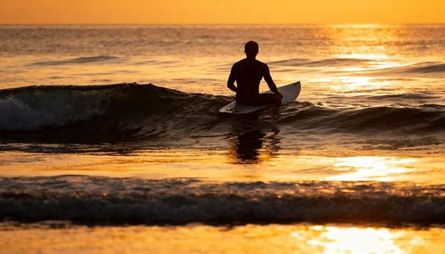 Solitary surfer sitting on surfboard facing golden sunset horizon over calm ocean waves