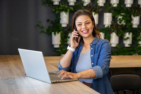 Smiling professional woman working on laptop taking phone call in coworking space