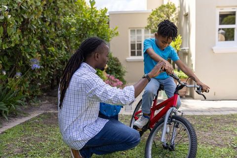 African american father guiding son learning to ride bicycle in suburban yard holding helmet
