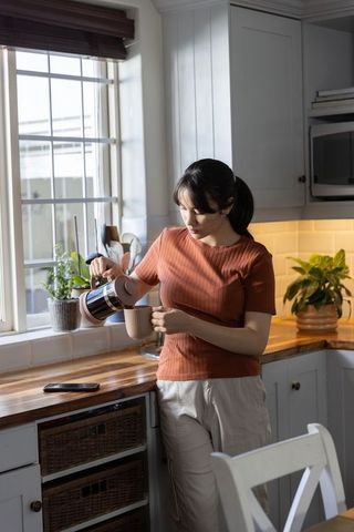 Young woman pouring coffee from stainless carafe into mug in sunlit cozy kitchen