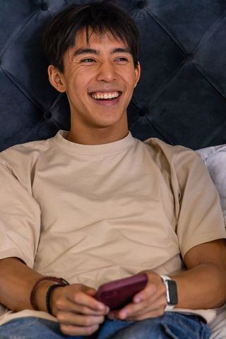 Smiling asian man relaxing on bed holding smartphone wearing smartwatch and beige t-shirt