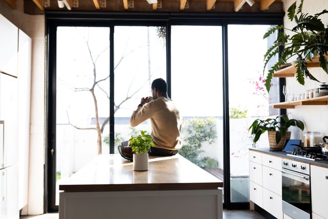 This image shows a man sitting on a kitchen counter, drinking coffee and looking out of large windows. The modern kitchen is filled with natural light and indoor plants, creating a peaceful and relaxing atmosphere. This image can be used for lifestyle blogs, articles about home decor, morning routines, or advertisements for kitchen appliances and home products.